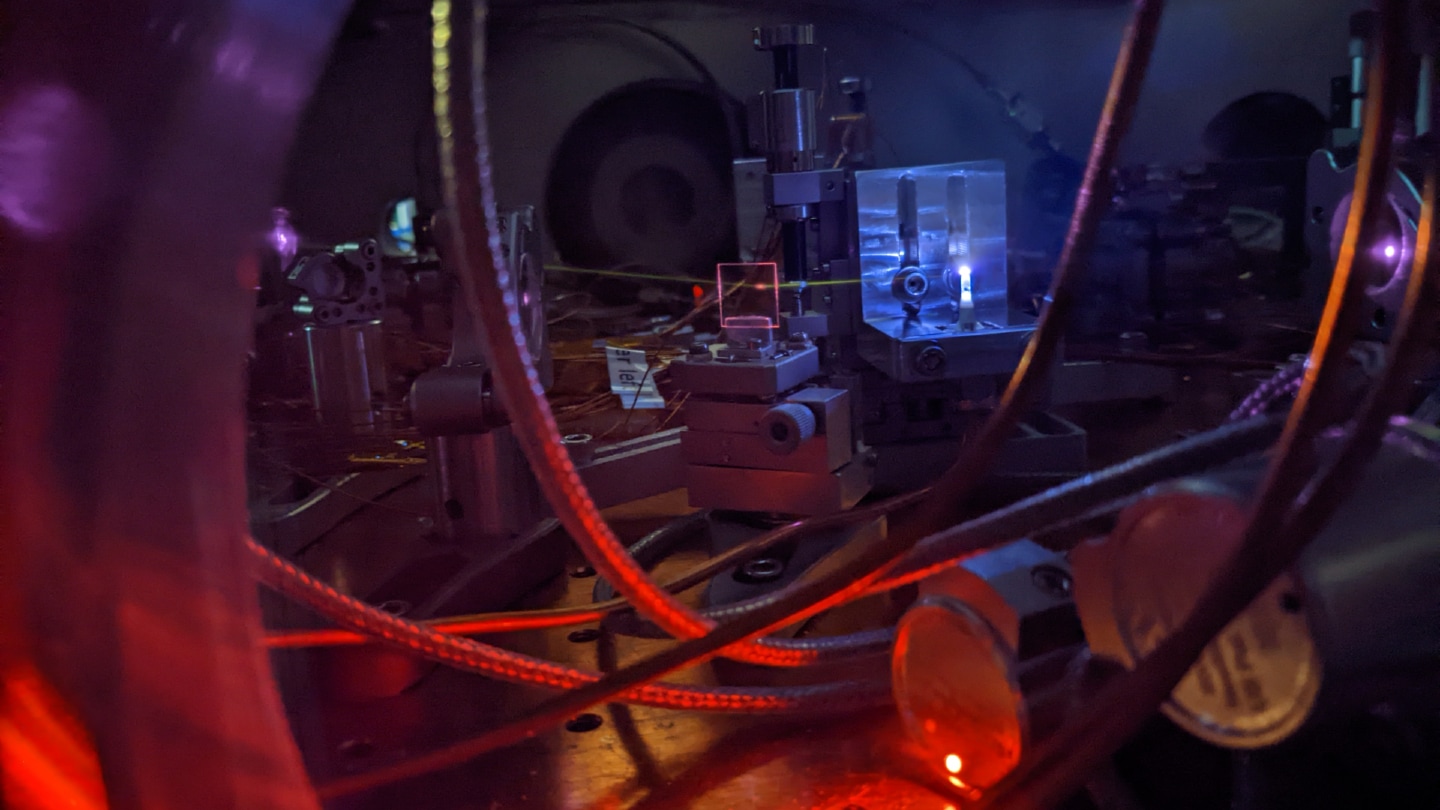 A photograph of scientific equipment, including a laser beam that illuminates gas inside a vacuum chamber.